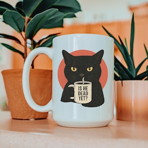 May include: White ceramic mug featuring a black cat illustration with yellow eyes and a red circle background. The cat holds a mug with the text "IS HE DEAD YET?" in black. The mug has a large handle.