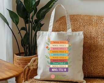 May include: Cream-colored tote bag featuring a stack of colorful books with the titles "NOVEL TITLE 1" through "NOVEL TITLE 10." Floral and leaf accents surround the book stack. The bag has two handles and is made of a sturdy fabric.
