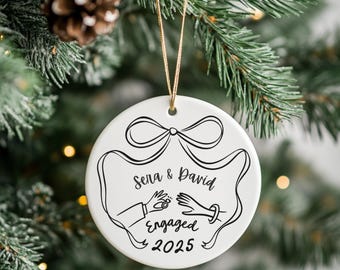 May include: White ceramic Christmas ornament with a black line drawing of two hands, a bow, and the words "Sena & David Engaged 2025." The ornament hangs from a gold string against a backdrop of a Christmas tree.
