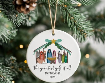 May include: White ceramic Christmas ornament with a Nativity scene illustration. The ornament features a stable with figures in colorful robes, a star, and the text "the greatest gift of all" with a Bible verse and year.