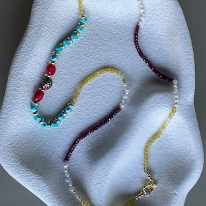May include: A multi-strand necklace featuring turquoise, red, and yellow beads, along with small white pearls. The necklace has a gold clasp and is displayed on a white, organic-shaped surface. The necklace is a piece of jewelry.