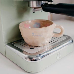 Puede incluir: Una pequeña taza de café de cerámica hecha a mano con un esmalte de color melocotón claro, apoyada en una bandeja de goteo plateada de una máquina de espresso verde menta. La taza está decorada con diseños florales rosas y azules y una palabra manuscrita en azul.