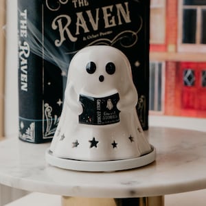 以下が含まれることがあります： 星の切り抜きと「Ghost Stories」と書かれた小さな本が付いた、白いセラミック製の幽霊型のキャンドルホルダー。幽霊は本を持っており、黒い目と口があります。「The Raven & Other Poems」というタイトルの本が背景にあります。
