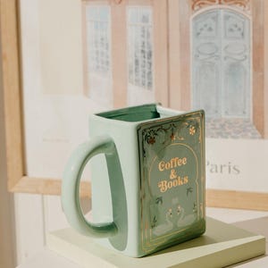 May include: A square, mint-green ceramic mug with a curved handle. The mug features gold lettering that reads "Coffee & Books" and a decorative swan design. The mug is displayed on a light green rectangular stand, with a framed picture in the background.