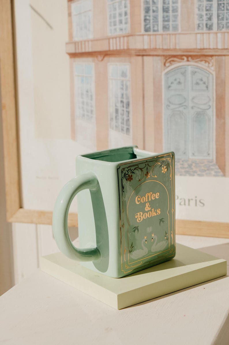 May include: A square, mint-green ceramic mug with a curved handle. The mug features gold lettering that reads "Coffee & Books" and a decorative swan design. The mug is displayed on a light green rectangular stand, with a framed picture in the background.