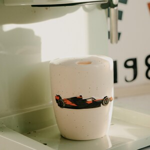 May include: A ceramic mug with a race car design in black and orange. The mug is white with small black speckles and sits on a light green tray. The mug is next to a white and green coffee machine.