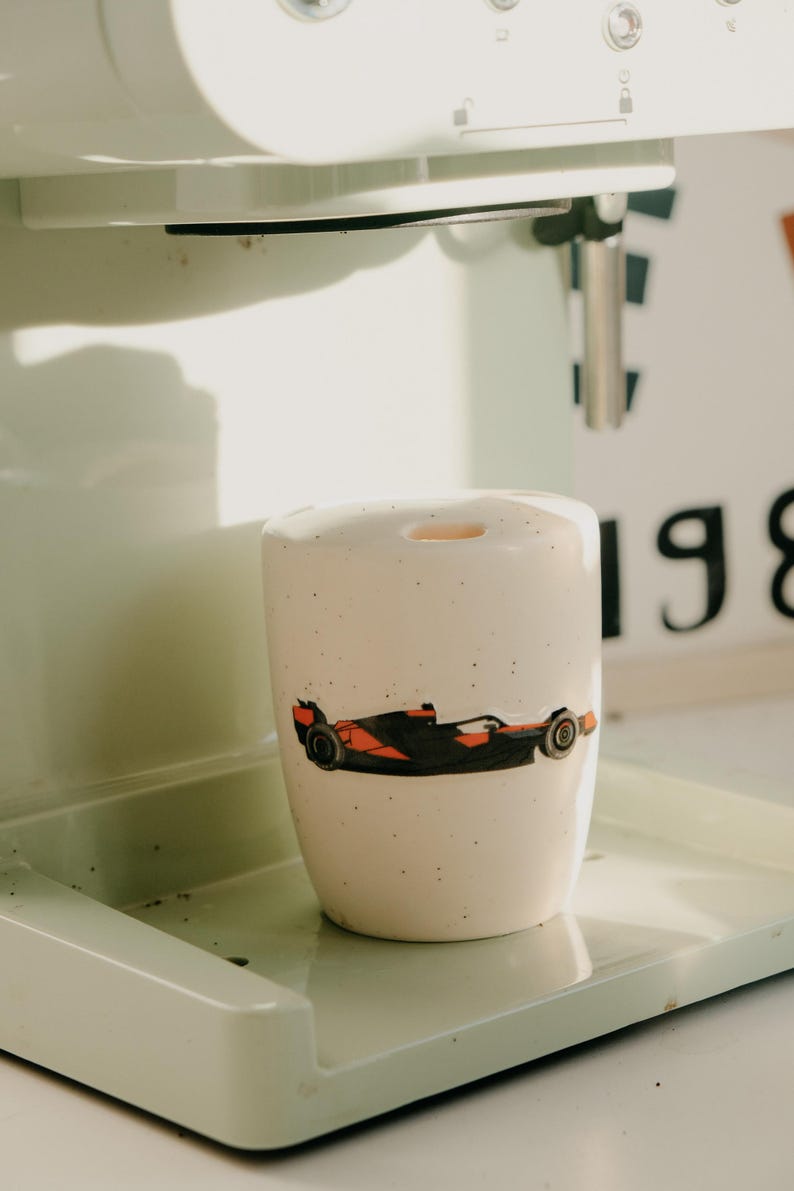 May include: A ceramic mug with a race car design in black and orange. The mug is white with small black speckles and sits on a light green tray. The mug is next to a white and green coffee machine.