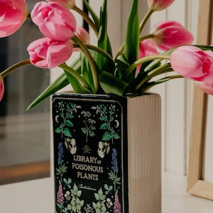 May include: A cream-coloured vase in the shape of a book, inscribed with "Library of Poisonous Plants", displays a bunch of pink tulips. The vase's front panel is black, decorated with botanical illustrations.