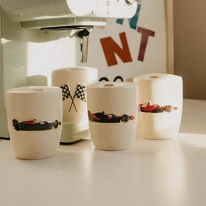 May include: Four off-white ceramic mugs with black, red, and orange race car designs. One mug has crossed checkered flags. The mugs are cylindrical and sit on a white surface, with a mint green appliance in the background.