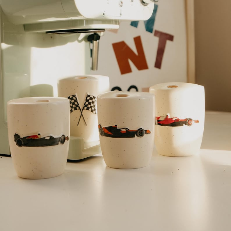 May include: Four off-white ceramic mugs with black, red, and orange race car designs. One mug has crossed checkered flags. The mugs are cylindrical and sit on a white surface, with a mint green appliance in the background.