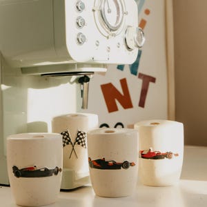 May include: A mint green coffee maker with a clear dial and chrome accents sits behind four cream-colored ceramic cups. Each cup features a race car design. One cup has crossed checkered flags.