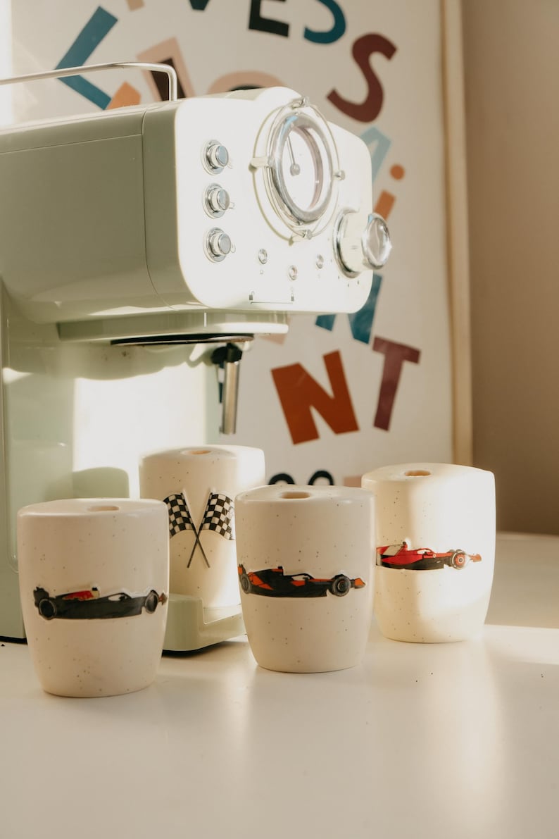 May include: A mint green coffee maker with a clear dial and chrome accents sits behind four cream-colored ceramic cups. Each cup features a race car design. One cup has crossed checkered flags.