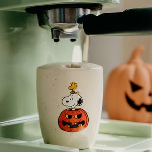 May include: A cream-colored ceramic mug with a speckled finish featuring a graphic of Snoopy and Woodstock sitting inside a carved orange jack-o'-lantern. The mug is positioned under the portafilter of a light green espresso machine.