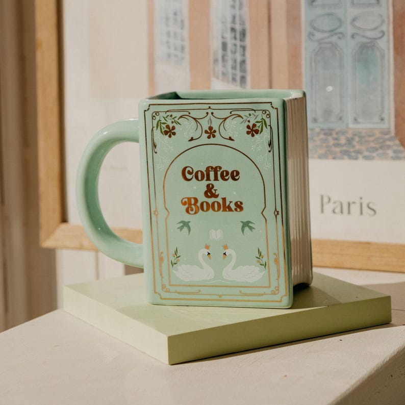 May include: A mint green ceramic mug shaped like a book, with the words "Coffee & Books" in gold lettering. The mug features a handle and a decorative design with swans and floral accents. The mug is sitting on a light green surface.