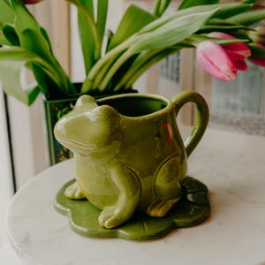 Op de afbeelding: Een groene keramische kikker-vormige mok met een handvat, op een groene blad-vormige schotel. De mok heeft een gedetailleerd kikkergezicht en poten. De achtergrond bevat een vaas met groene bladeren en roze tulpen.