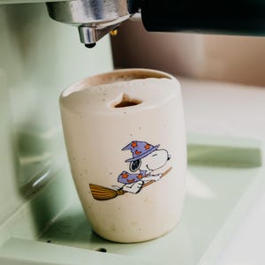 May include: A cream-colored ceramic mug with a speckled finish featuring a graphic of Snoopy wearing a purple wizard hat and cape while riding a broomstick. The mug has a unique top opening and is positioned under the portafilter of a light green espresso machine.