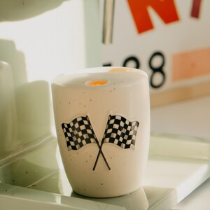 May include: A white ceramic cup with two crossed black and white checkered flags. The cup is speckled with small black dots and sits on a light green surface. The background includes a blurred sign with orange and red letters.