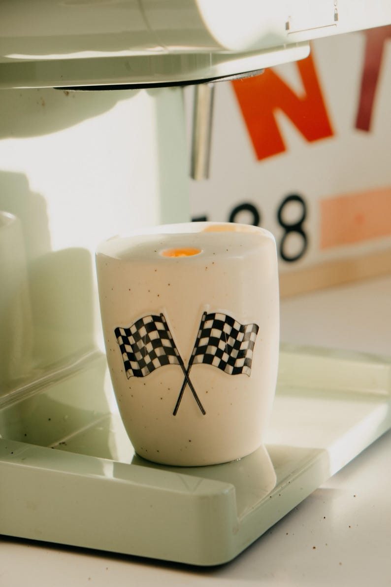 May include: A white ceramic cup with two crossed black and white checkered flags. The cup is speckled with small black dots and sits on a light green surface. The background includes a blurred sign with orange and red letters.
