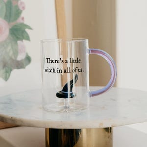 May include: A clear glass mug with a black witch hat design and the text "There's a little witch in all of us." The mug has a purple and blue iridescent handle. The mug is on a white marble surface.