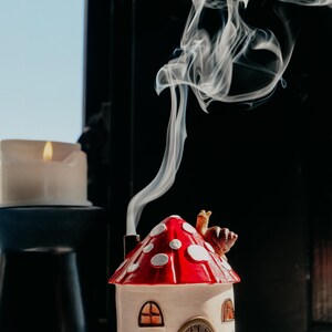 Pode incluir: Um queimador de incenso em forma de cogumelo com um telhado vermelho e branco com bolinhas. A casa de cer&acirc;mica tem uma pequena chamin&eacute; que emite fumo. Uma vela acesa est&aacute; por perto. O queimador est&aacute; sobre uma base azul.