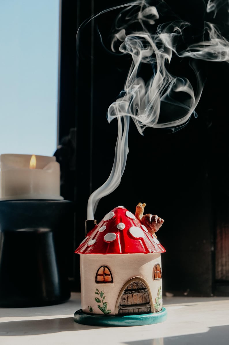 Pode incluir: Um queimador de incenso em forma de cogumelo com um telhado vermelho e branco com bolinhas. A casa de cer&acirc;mica tem uma pequena chamin&eacute; que emite fumo. Uma vela acesa est&aacute; por perto. O queimador est&aacute; sobre uma base azul.