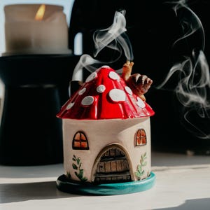 May include: A ceramic mushroom house incense burner with a red cap and white spots. The house has a beige body, arched doorway, and two arched windows. Smoke rises from the chimney, and a snail figurine sits on top. The burner sits on a teal base.