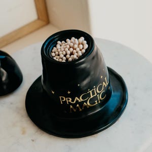 May include: A black ceramic match holder in the shape of a top hat, filled with matches. The words "PRACTICAL MAGIC" are printed in gold on the side of the hat, with small star accents. The holder sits on a round base.
