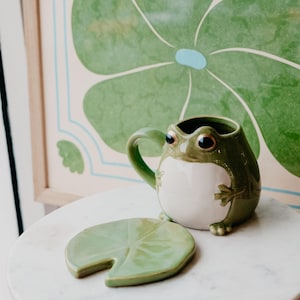 May include: A green ceramic frog mug with a white belly and a green leaf-shaped coaster. The mug has a handle and large eyes. The coaster is shaped like a lily pad.