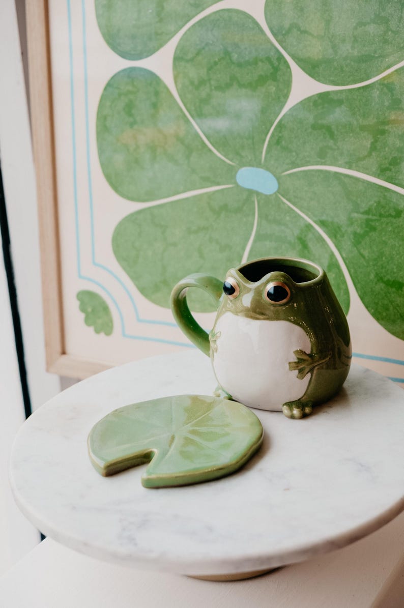 May include: A green ceramic frog mug with a white belly and a green leaf-shaped coaster. The mug has a handle and large eyes. The coaster is shaped like a lily pad.