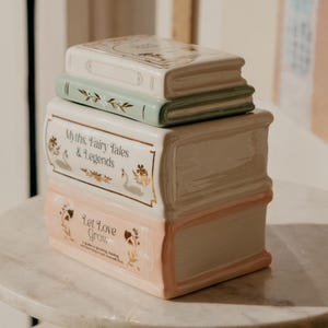 Peut inclure: Une pile de boîtes en céramique en forme de livres, de couleur crème, vert pâle et rose. Les boîtes sont décorées d'accents dorés, de motifs floraux et des phrases "Myths, Fairy Tales & Legends" et "Let Love Grow".