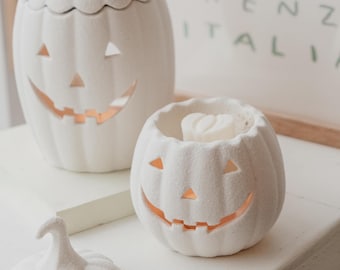 May include: Two white ceramic pumpkin-shaped candle warmers with a carved jack-o'-lantern face. The smaller warmer has a white wax melt inside.