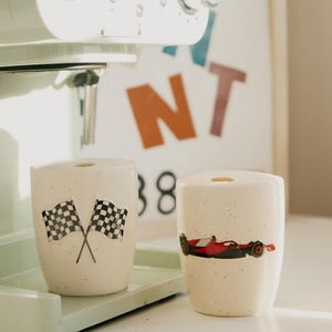 May include: Two speckled ceramic mugs with racing-themed designs. One mug features crossed checkered flags, and the other displays a red and black race car. The mugs sit on a light green tray, near a coffee machine.