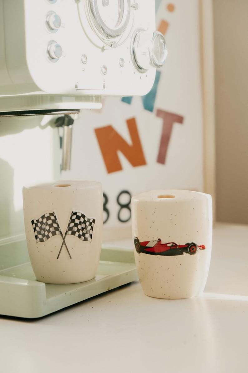 May include: Two speckled ceramic mugs with racing-themed designs. One mug features crossed checkered flags, and the other displays a red and black race car. The mugs sit on a light green tray, near a coffee machine.