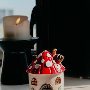 Pode incluir: Uma figura de casa de cogumelos caprichosa com uma tampa vermelha salpicada de manchas brancas. A casa tem um corpo bege, janelas arqueadas e uma porta de madeira. Uma pequena figura de caracol est&aacute; no topo do cogumelo. Uma vela acesa ao fundo.