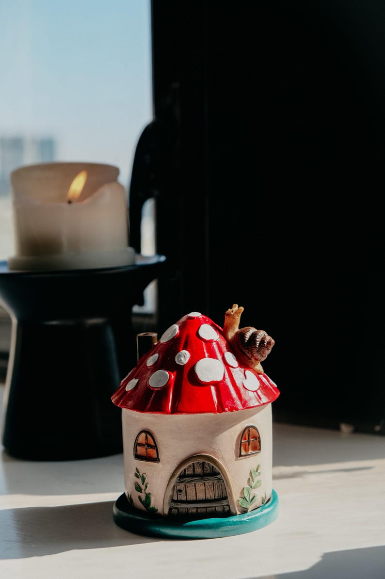 Pode incluir: Uma figura de casa de cogumelos caprichosa com uma tampa vermelha salpicada de manchas brancas. A casa tem um corpo bege, janelas arqueadas e uma porta de madeira. Uma pequena figura de caracol est&aacute; no topo do cogumelo. Uma vela acesa ao fundo.