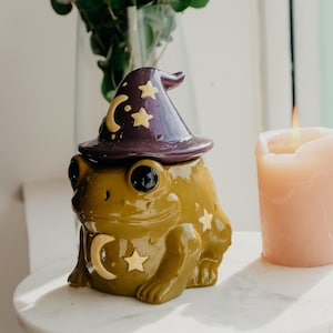 Op de afbeelding: Een keramische pot in de vorm van een kikker met een glanzende olijfgroene afwerking, voorzien van een afneembaar donkerpaars heksenhoeddeksel versierd met goudkleurige halve maan- en stermotieven op het lichaam van de kikker.