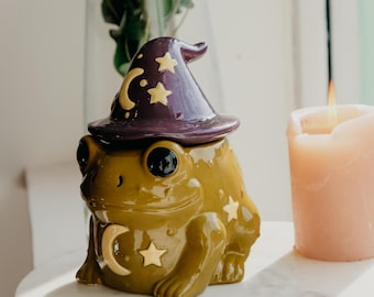 May include: A ceramic frog-shaped jar with a glossy olive green finish, featuring a removable dark purple witch hat lid decorated with gold-colored crescent moon and star motifs. The frog body also displays matching gold moon and star accents.