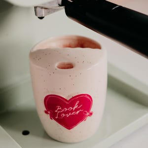 May include: A speckled, off-white ceramic mug with a red heart graphic that reads "Book Lover." The mug is inside a mint green espresso machine. The mug has a small hole at the top and is filled with a light pink liquid.