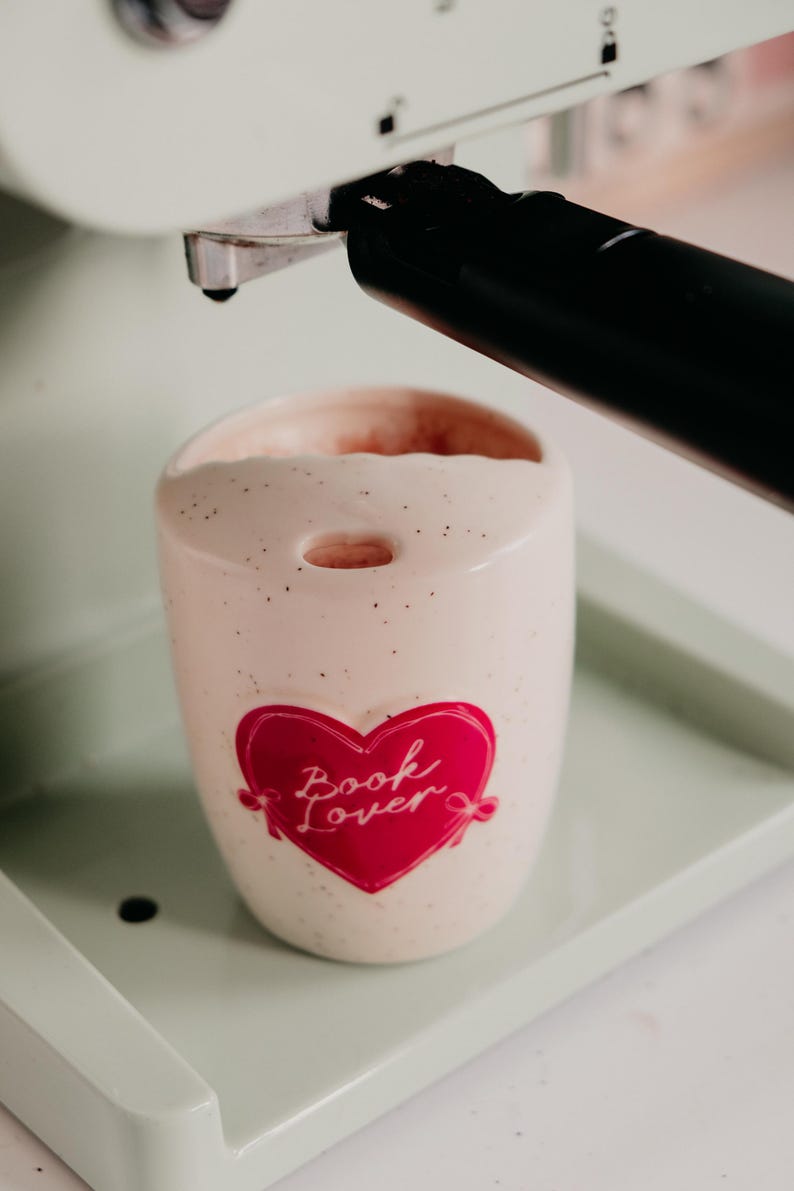 May include: A speckled, off-white ceramic mug with a red heart graphic that reads "Book Lover." The mug is inside a mint green espresso machine. The mug has a small hole at the top and is filled with a light pink liquid.