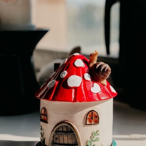 Pode incluir: Uma casa de cogumelos de cer&acirc;mica com um chap&eacute;u vermelho com manchas brancas e um caracol no topo. A casa tem janelas arqueadas e uma porta de madeira, assente numa base azul-turquesa. Folhas decorativas adornam a lateral.