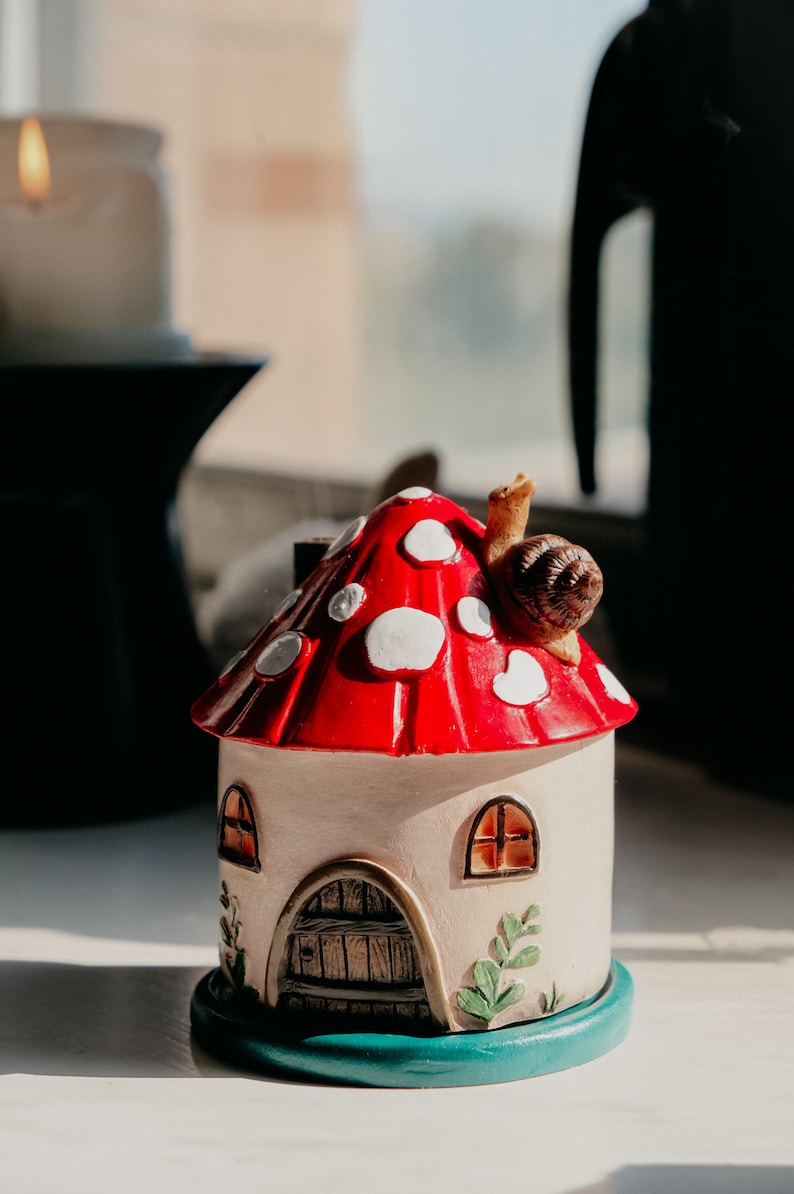 Pode incluir: Uma casa de cogumelos de cer&acirc;mica com um chap&eacute;u vermelho com manchas brancas e um caracol no topo. A casa tem janelas arqueadas e uma porta de madeira, assente numa base azul-turquesa. Folhas decorativas adornam a lateral.