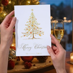 Può includere: Un biglietto di Natale bianco con un'illustrazione dorata di un albero di Natale, adornato con ornamenti dorati e arancioni. Il biglietto recita "Merry Christmas" in elegante scrittura. Il biglietto è tenuto davanti a un albero di Natale e decorazioni festive.