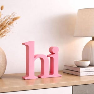 Puede incluir: Letras rosas de madera que deletrean "hi" se exhiben sobre una superficie de madera. A la izquierda hay un jarr&oacute;n beige con flores secas, y a la derecha una l&aacute;mpara blanca y libros. Las letras son un acento decorativo.