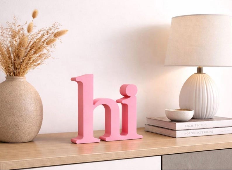 Puede incluir: Letras rosas de madera que deletrean "hi" se exhiben sobre una superficie de madera. A la izquierda hay un jarr&oacute;n beige con flores secas, y a la derecha una l&aacute;mpara blanca y libros. Las letras son un acento decorativo.