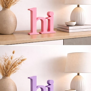 Puede incluir: Letras "hi" de madera rosa sobre una estanter&iacute;a de madera, con un jarr&oacute;n de flores secas, una l&aacute;mpara y libros. Las letras miden unos 15 cm de alto. La segunda imagen muestra las mismas letras en morado.