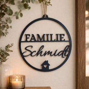Puede incluir: Un letrero familiar de madera azul oscuro con la palabra "FAMILIE" sobre el nombre "Schmidt" en escritura cursiva. Una pequeña casa con un corazón recortado está en la parte inferior. El letrero circular cuelga de una cuerda natural.