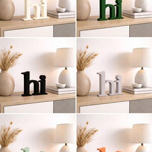 Puede incluir: Una colecci&oacute;n de letreros decorativos de madera con la palabra "hi" en varios colores, incluyendo blanco, verde, negro, gris, verde menta y naranja. Los letreros se exhiben sobre una superficie de madera clara, junto a jarrones, l&aacute;mparas y libros.