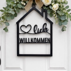 Puede incluir: Un letrero de madera negro en forma de casa con la palabra "Willkommen" y un símbolo de corazón, colgado en una puerta blanca. El letrero está decorado con vegetación, rosas blancas y un lazo de arpillera.