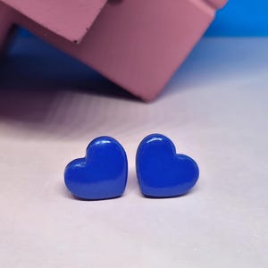 Puede incluir: Dos pendientes de botón en forma de corazón azul cobalto brillante. Los pendientes están colocados sobre una superficie blanca, con un fondo rosa y azul. Un diseño sencillo y elegante.