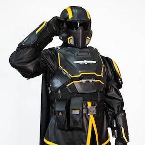 May include: Black and yellow futuristic armor with a helmet, chest plate, and leg guards. The armor has yellow accents and a skull emblem. The person is wearing black gloves and a black cape. The overall design suggests a sci-fi or cosplay theme.